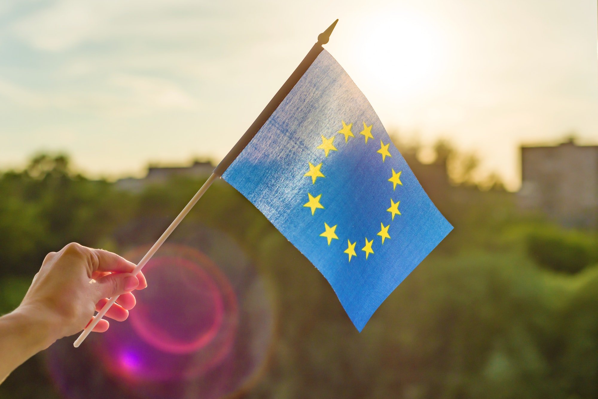 Hand holds European Union flag in an open window. Background blue sky, silhouette of the city